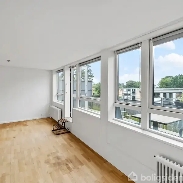 Et tomt værelse har parketgulv, store vinduer langs den ene væg, der giver udsigt til et boligområde med himmel og træer i baggrunden. Ingen tekst er til stede.