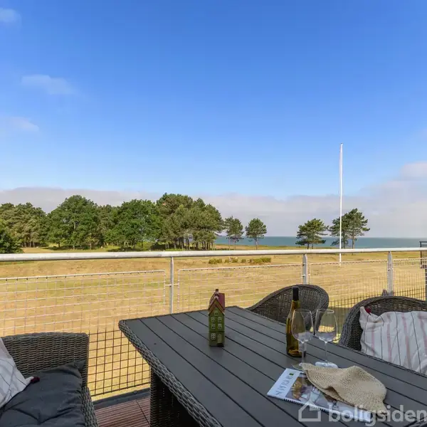 Terrasse med bord og stole arrangeret til afslapning. Bordet har en flaske vin og glas. Udsigt over græsplæne, træer og havet i baggrunden. Klar himmel.