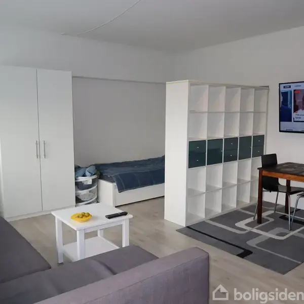 Et komfur med elkedel og kogeplader står i et køkken med hvidt skab. Belysning er tændt under overskabene. I baggrunden ses en stue med en sofa og spisebord.