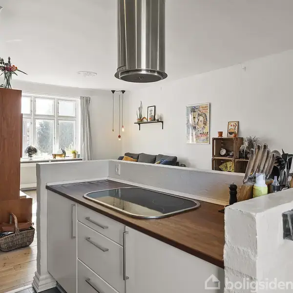 Køkkenområde med vask og ovn i en ø, træbordplade, omgivet af stue med sofa, vindue med dagslys, hylder og dekorationer på væggene. Kaffeartikler og knive er på bordet.