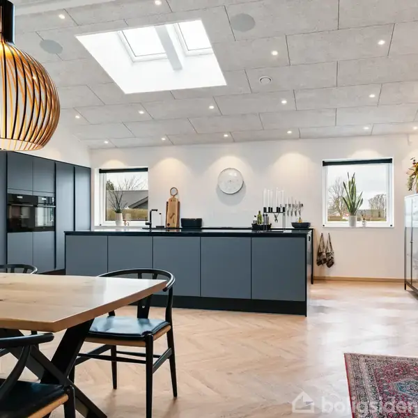 Et moderne køkken med mørke skabe står i centrum, omgivet af en stue med trægulv og murstensvægge. Et stort spisebord og loftsvindue skaber en lys, rummelig atmosfære.