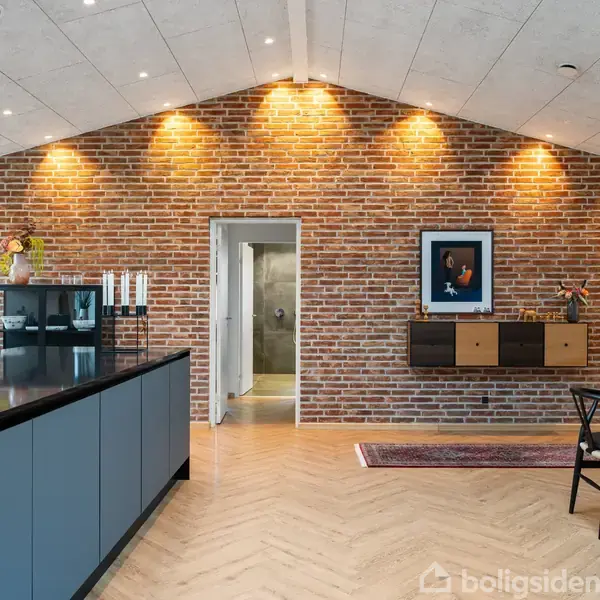 Værelsesinteriør med murstensvæg, hvor der hænger et billede og en hylde. En køkkenø med sorte overflader ses til venstre. Trægulv og spotsbelysning skaber en hyggelig atmosfære.