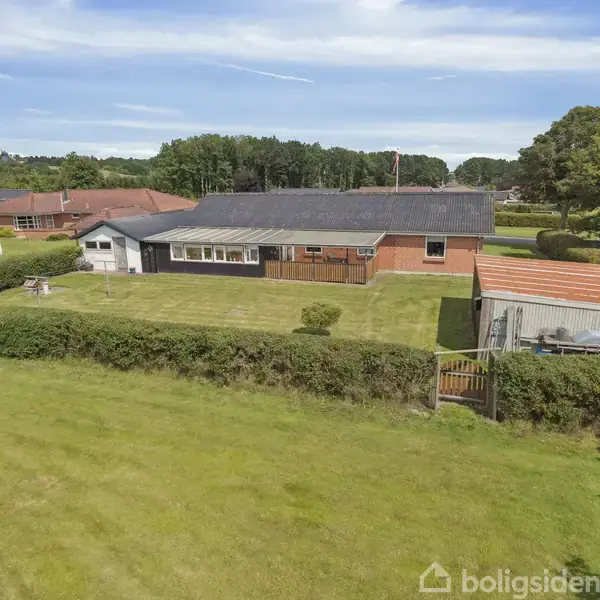 Et enkelt familiehus i røde mursten står midt i en grøn have omkranset af lave hække. En garage og skur ses ved siden af, omgivet af græs og træer.