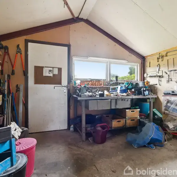 Værksted med rodet arbejdsbord fuld af værktøj, omgivet af redskaber hængt på væggen. Der er en dør og et vindue med udsigt. Hylder langs væggen opbevarer diverse kasser.