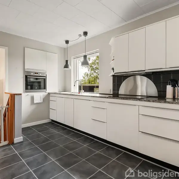 Hvidt køkken med sorte gulvfliser indeholder lange bordplader, indbyggede ovne og moderne lamper. En trappe fører nedad ved siden af køkkenet, der har åbne hylder med porcelæn.