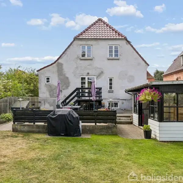 Hvidt hus med rødt tag står bag en terrasse med sort hegn. Grill dækket af sort betræk ses foran, omgivet af græs og have.