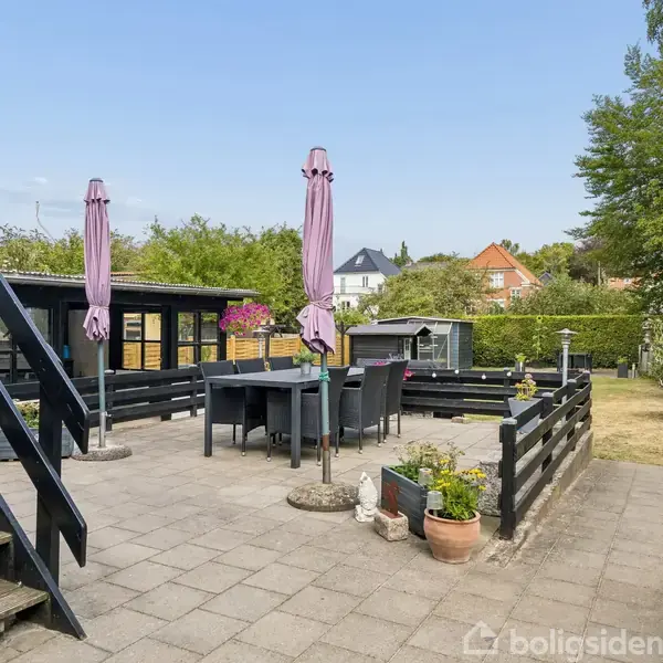 En udendørs terrasse med et sort bord og stole under lyserøde parasoller. Trappen fører op fra terrassen til et grønt haverum omkranset af træer og buske.