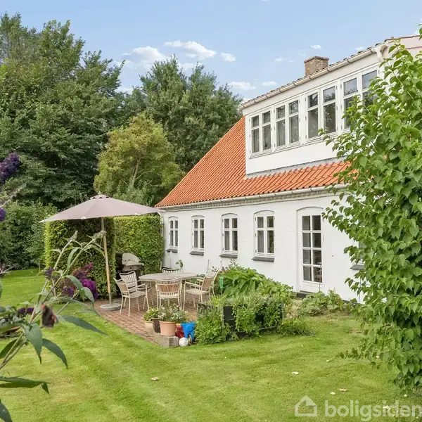 Hvidt hus med rødt tag står i sollys. Terrasse med havemøbler og parasol er omgivet af grønne buske og træer. Blomster pryder forhaven, skaber en hyggelig haveatmosfære.