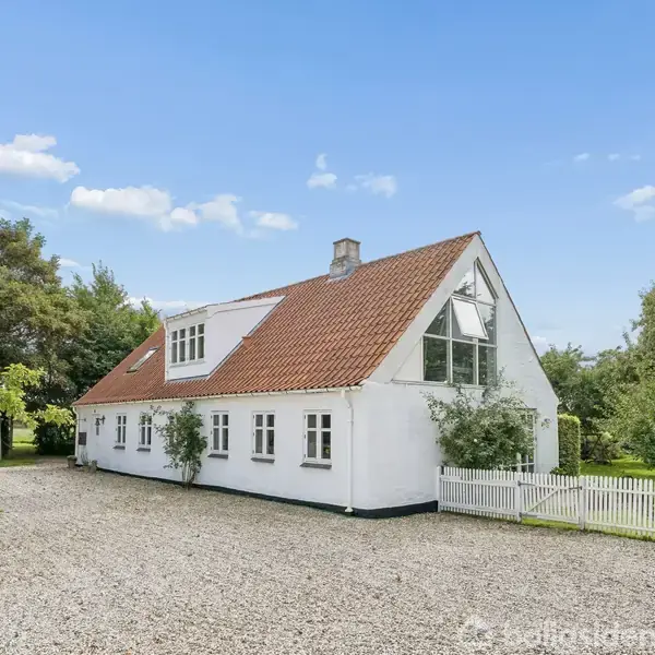 En hvidkalket landlig ejendom med rødt tegltag står i en grøn have. Ejendommen har mange vinduer og en grusbelagt indkørsel omgivet af træer i solrigt vejr.