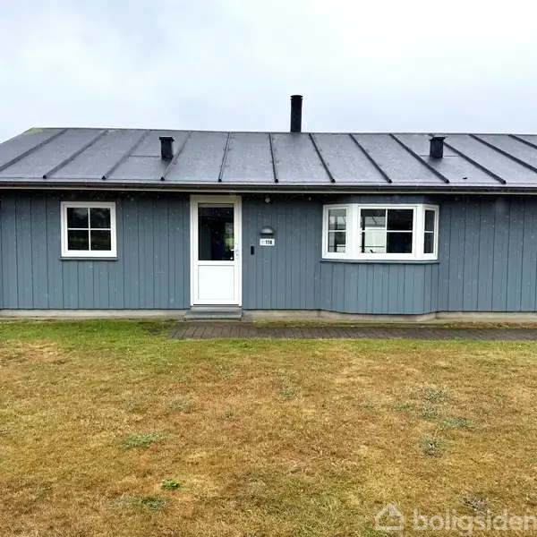 Blåmalet træhus står stille på en græsplæne under overskyet himmel. Huset har to vinduer og en hvid dør i midten, markeret med "A10".