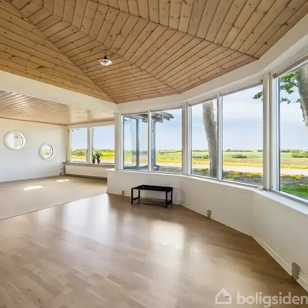 Lyst rum med trægulv og træloft har store panoramavinduer med udsigt over en grøn mark og himmel. En bænk er placeret foran vinduerne.