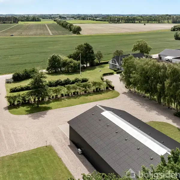 En stor ladebygning med sort tag ligger på en grusvej omgivet af træer og grønne marker i et landdistrikt med et hus i baggrunden.