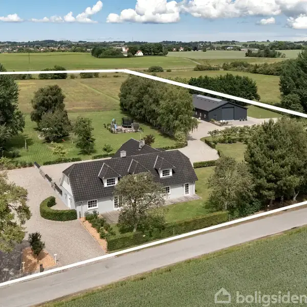Hus med sort tag står blandt træer og grønt græs, indrammet af hvid linje. Lang vej i forgrunden, marker strækker sig i baggrunden under blå himmel med skyer.