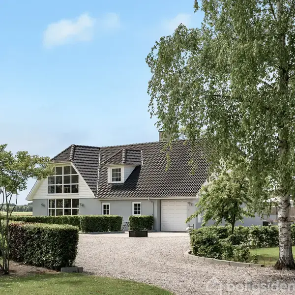 Et moderne hus med en mørk tag, omgivet af en velplejet have med træer og hække, står i et grønt landskab under en klar blå himmel.
