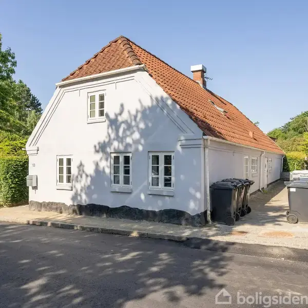 Hvidt hus med rødt tag står stille på hjørnet af en vej. Skraldespande står ved siden. Træer skaber skygger på facaden i et grønt boligområde.