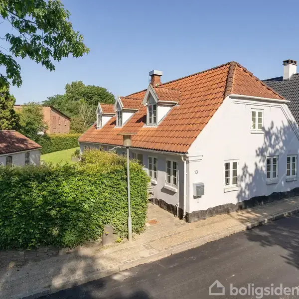 Hvidmalet hus med rødt tegltag og flere kviste, beliggende ved en gade med fortov samt grønne buske i baggrunden. Skygger fra træerne falder på vejen.