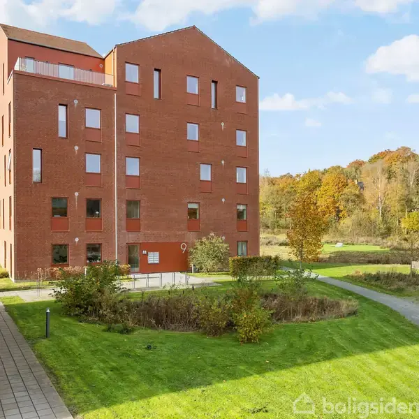 En rød murstensbygning med flere etager står i et grønt område. Gehveje og stier omgiver bygningen, mens træer og buske viser en naturlig baggrund.