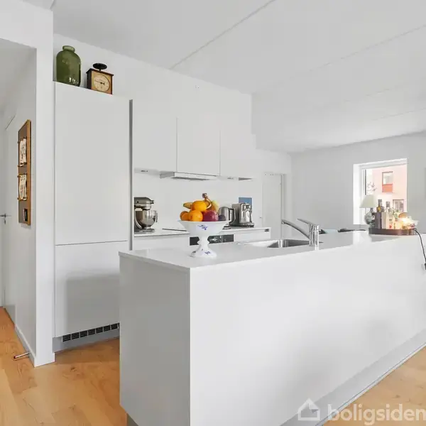 Et moderne køkken med en hvid ø står midt i et rum med trægulv. Frugt og køkkenmaskiner er placeret på øen. En sofa er synlig i baggrunden.