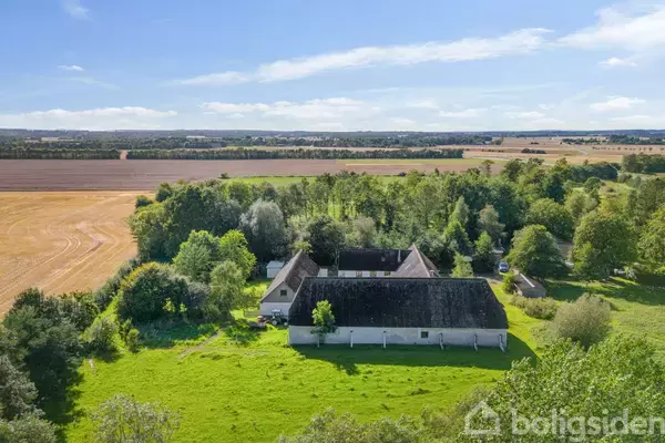 En hvid gård med stråtag ligger omgivet af grønne marker og skove. Den rolige landlige scene strækker sig mod en vid horisont under en klar blå himmel.