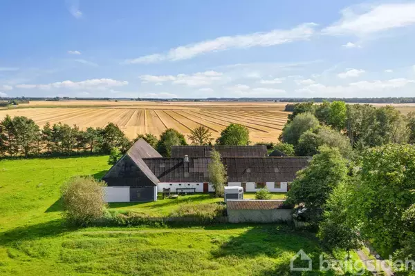 En landlig gård med hvide bygninger og mørkt tag ligger omgivet af frodige, grønne træer. I baggrunden strækker bølgende gulbrune marker sig under en klar blå himmel.