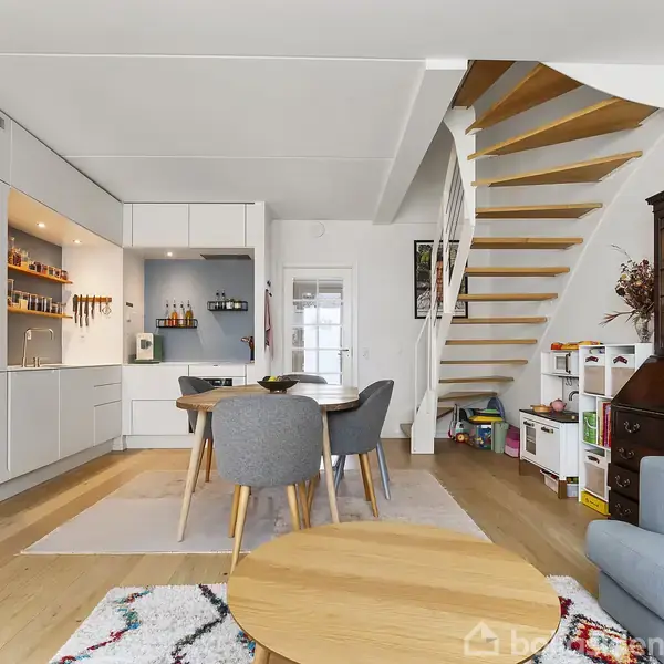 Spisebord med stole står i en moderne, åben stue med køkken i baggrunden. En åben trætrappe fører til et andet niveau. Hylder og skabe fylder væggene.