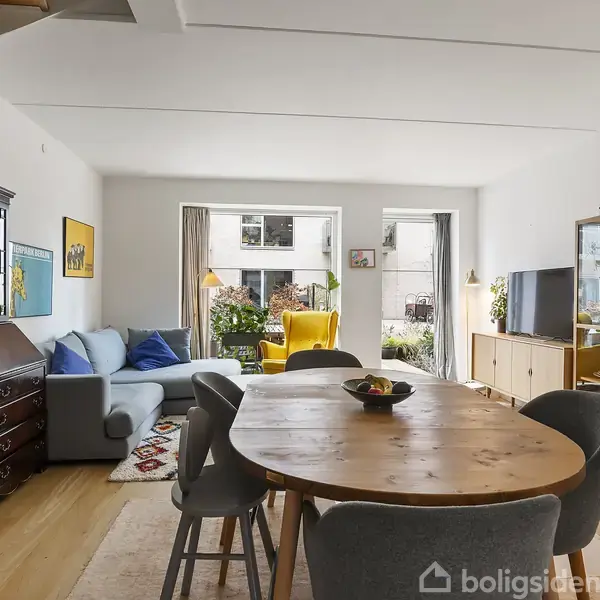 Et rundt træbord med stole er centralt placeret i en lys stue. Rummet har en blå sofa, gul lænestol, reoler og vinduer, der giver udsigt til terrasse og have.