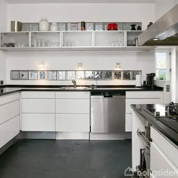 Et moderne køkken med hvide skabe. En opvaskemaskine er indbygget under sort bordplade. Hylder med glasåbninger over vasken. Væggene har kunstværker, og der er en stor kogeø.