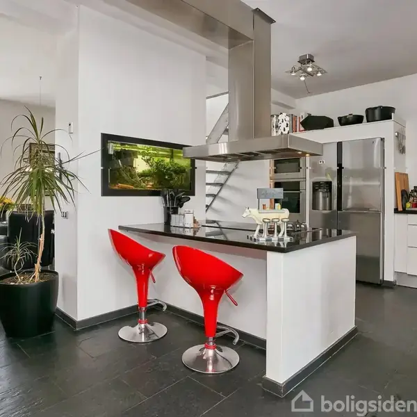 Køkkenø med to røde barstole står ubenyttet i moderne køkken. Omgivet af hvidvægge, stor plante og spisestue i baggrunden. Blik mod åben ruminddeling med grå gulvfliser.