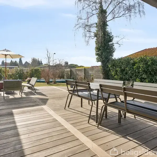 Terrassebord og stole står på en træterrasse. Parasol giver skygge over andre udendørsmøbler. Omkring er en frodig have med buske og træer. Klar blå himmel ses ovenover.