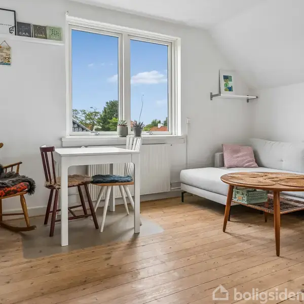 Stue med træbord og hvid sofa ved siden af et mindre spisebord med to stole. Lyse omgivelser gennem et stort vindue og grønne planter på gulvet.