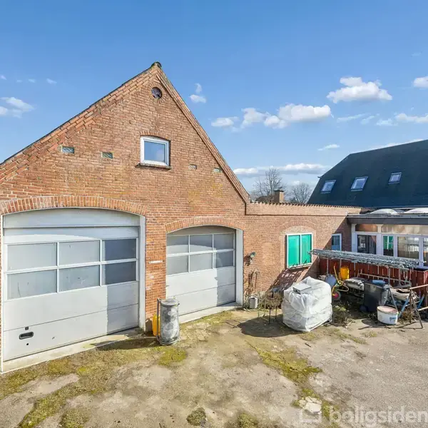 Rødt murstenshus med to store garageporte står i solen. Omkring huset ses en gårdsplads med diverse udstyr og beholdere. I baggrunden er der et område med træer og himmel.