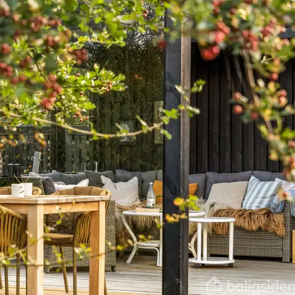 Skydækket terrasse med havemøbler, herunder en sofa med puder og et bord med stole. Planter med røde bær omkranser terrassen, og en sort lanterne hænger fra loftet.