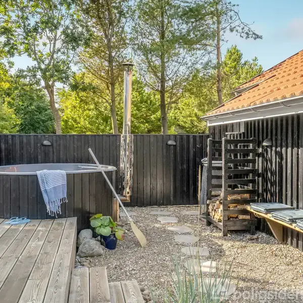 Brunt vildmarksbad står på en træterrasse. Et tæppe og en åre hviler på kanten. Baggrunden har et sort hegn, et skur med rødt tag og omgivelser af frodig skov.