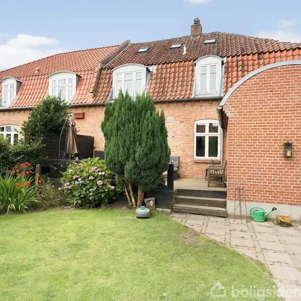 Et rødt murstenshus med rødt tegltag har en velplejet have med blomster og en terrasse. Huset har skråvinduer på første sal og flere trapper, der fører op til dets indgang.