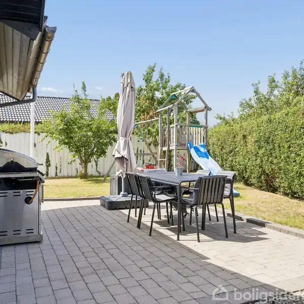Grill og spisebord står på terrasse med fliser i en have. Der er en parasol ved bordet og et legeområde med rutsjebane i græsset, omgivet af buske.