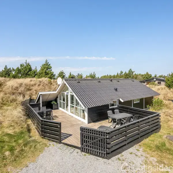 Et sort sommerhus med stort vinduesparti står på en terrasse omgivet af træhegn. Det ligger i et naturlandskab af græs og fyrretræer under en blå himmel.