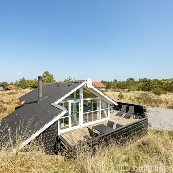 Sommerhus med stort glasparti står tomt; omgivet af græsklædte klitter og sparsom bebyggelse under en klar, blå himmel. Terrasse med møbler fuldender det fredfyldte, naturskønne miljø.