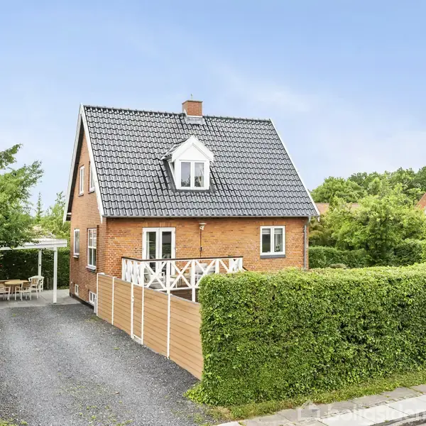 Rødstenshus med sort tag står stille ved en grusindkørsel. Til højre er en terrasse med hvidt stakit, omgivet af frodige grønne buske og træer.