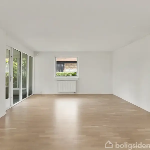 Et rummeligt værelse med lyst trægulv, hvide vægge og store vinduer. Vinduet foran viser udsigt til en have. Til højre er der en åbning til et andet rum.