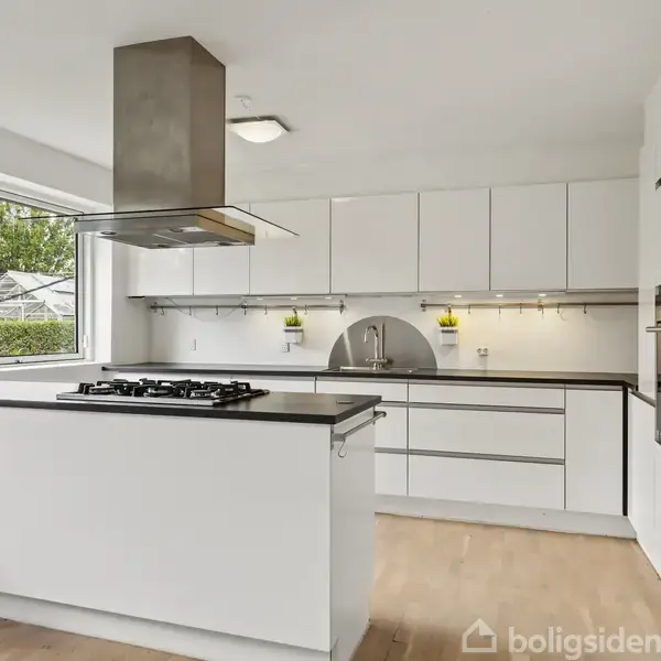 Et moderne køkken med en kogeø, udstyret med gaskomfur og emhætte, er omgivet af hvide skabe. Store vinduer giver naturligt lys, med udsigt til havegrønne. Trægulve supplerer rummet.