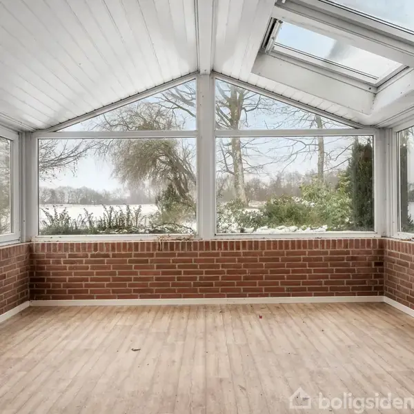 Udestue med store vinduer og loftsvinduer slipper vinterlys ind. Omgivet af snedækket landskab og træer uden for. Røde murstensvægge og trægulv dominerer rummet.