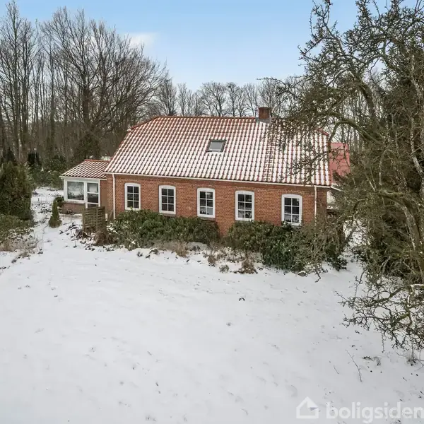 Et rødt murstenshus med et rødt tegltag står stille i et snedækket landskab, omgivet af træer og buske i en vinteragtig have.