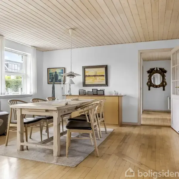 Spisebord med seks træstole står på et tæppe i en lys stue med trægulv og hvide vægge. Væggen har to malerier, og der hænger en pendellampe over bordet.
