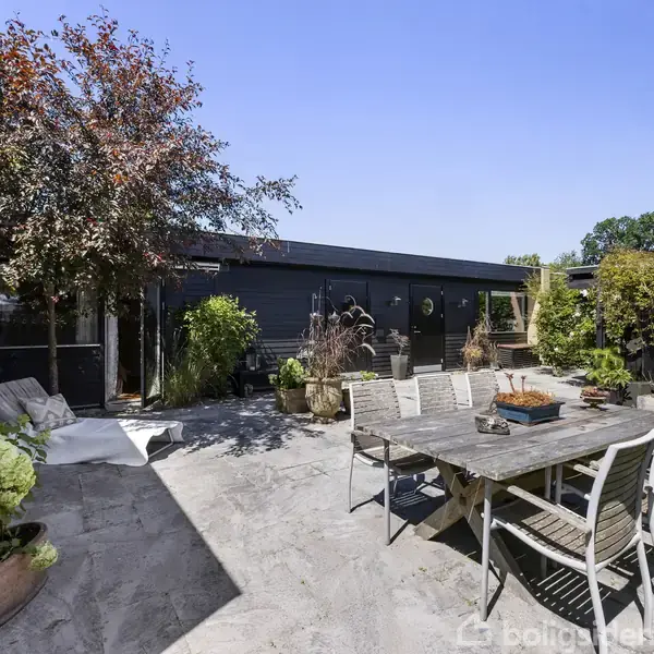 Udendørs spisebord med stole står på en flisebelagt terrasse omgivet af grønne planter og en træmøbleret bygning i baggrunden under en klar blå himmel.