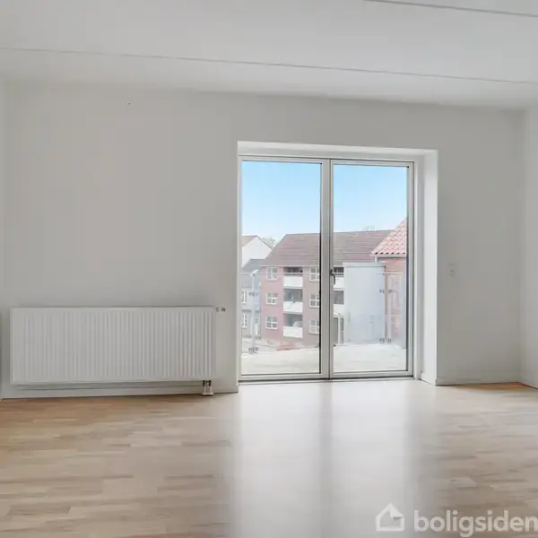 Et lyst rum med trægulv, hvor en radiator står mod venstre væg. En glasdør i centrum leder ud til en balkon med udsigt over omkringliggende bygninger.