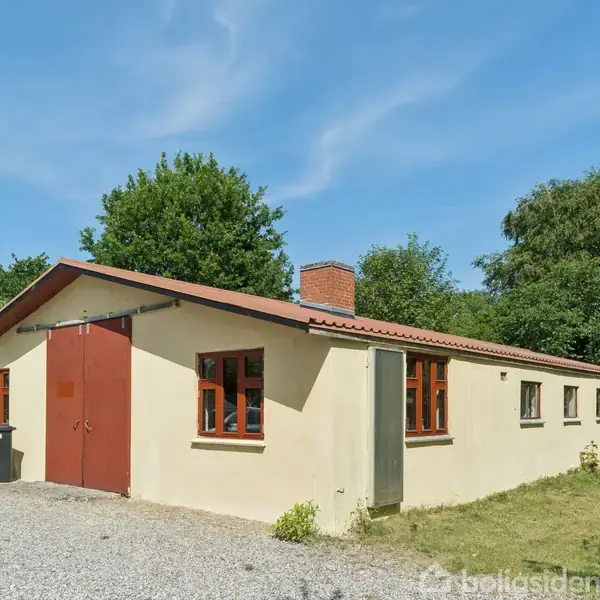 En lang, étplans gul bygning med rødt tag og døre. Bygningen står på en grusbelagt plads omgivet af græs og træer under en klar blå himmel.