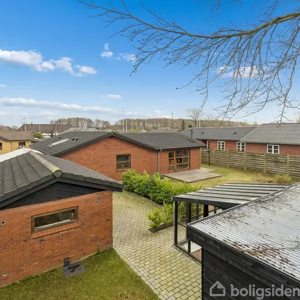 Rødt murstenshus med sort tag står i en indhegnet have med flisebelægning. Omgivet af flere huse under en blå himmel med spredte skyer.