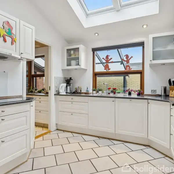 Et lyst køkken med hvide skabe, dekoreret med farverige figurer. Rummet har en ovn, kaffemaskine og store vinduer, der giver naturligt lys fra ovenlysvinduer. Flisegulv bidrager til det moderne udtryk.