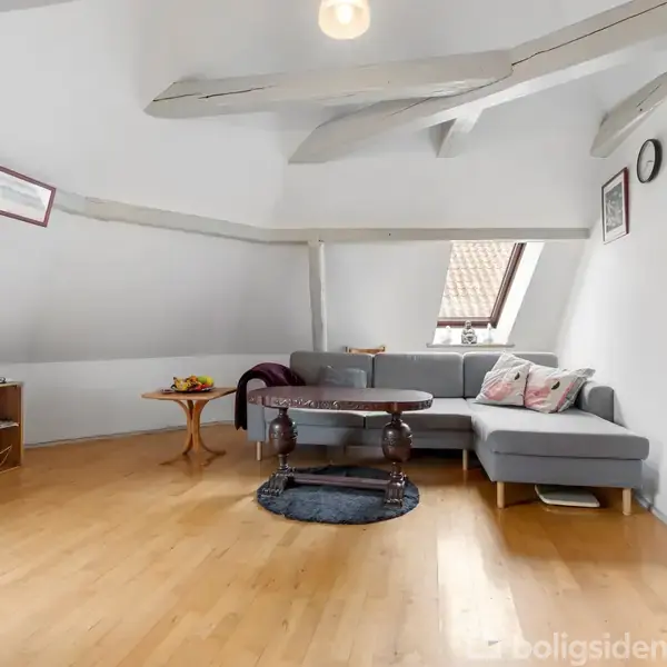 Grå sofa står i en stue med skråvægge og trælofter. Et rundt sofabord er placeret på et lille rundt tæppe. Hylder med bøger ses til venstre.