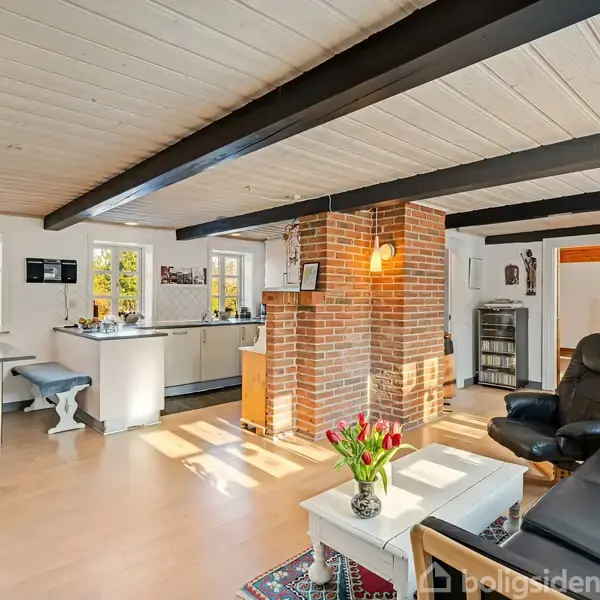 Stue med sofa og lænestol ved siden af et kaffebord med tulipaner, adskilt fra køkkenet af en murstensskorsten. Væggene er hvide, og rummet er lyst op af vinduer.